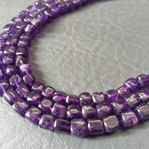 Puede incluir: Una hebra de cuentas de amatista púrpura, cada cuenta está facetada y tiene forma cuadrada.