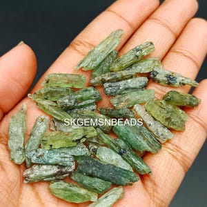 May include: A collection of green and black gemstone crystals, known as actinolite, in various sizes and shapes. The crystals are raw and have a natural, rough texture. The crystals are displayed in a hand, showcasing their unique beauty.