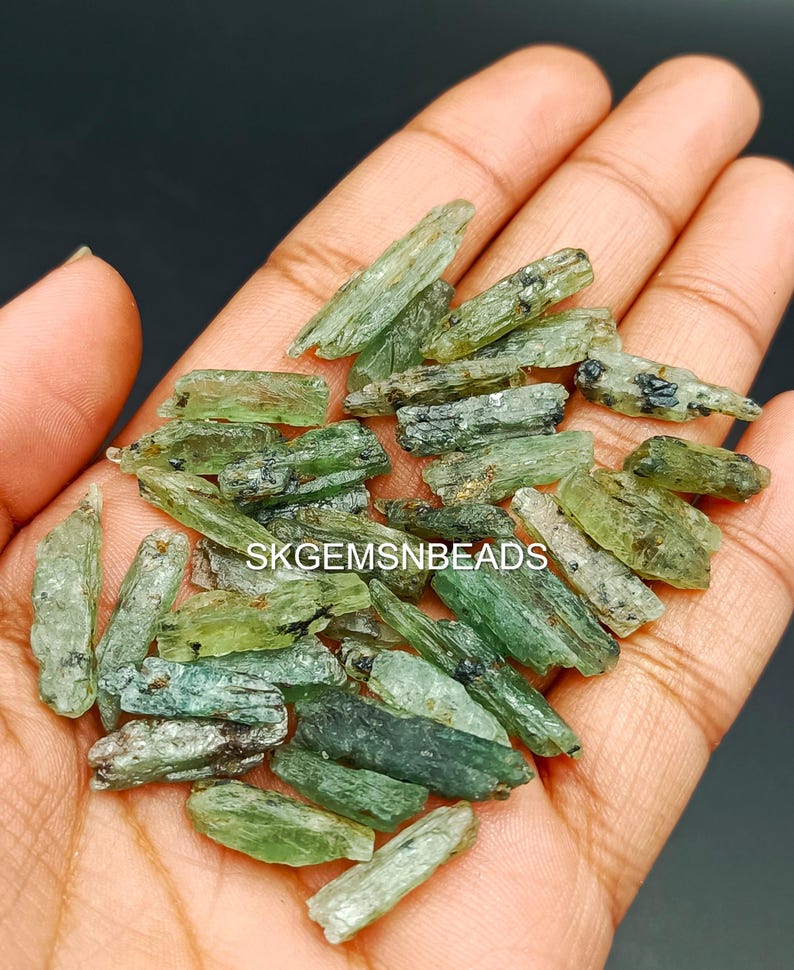 May include: A collection of green and black gemstone crystals, known as actinolite, in various sizes and shapes. The crystals are raw and have a natural, rough texture. The crystals are displayed in a hand, showcasing their unique beauty.