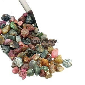 K&ouml;nnte beinhalten: Eine Schaufel mit bunten rohen Edelsteinen, darunter rosa, gr&uuml;n, blau und braun, in verschiedenen Gr&ouml;&szlig;en und Formen. Die Edelsteine sind auf einer wei&szlig;en Oberfl&auml;che verstreut.