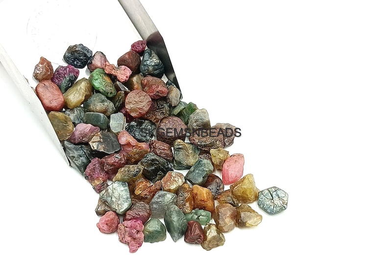 K&ouml;nnte beinhalten: Eine Schaufel mit bunten rohen Edelsteinen, darunter rosa, gr&uuml;n, blau und braun, in verschiedenen Gr&ouml;&szlig;en und Formen. Die Edelsteine sind auf einer wei&szlig;en Oberfl&auml;che verstreut.