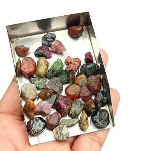 K&ouml;nnte beinhalten: Eine Sammlung roher, ungeschliffener Edelsteine in verschiedenen Farben, darunter Rot, Gr&uuml;n, Blau und Schwarz, die in einer kleinen Metallschale pr&auml;sentiert werden.