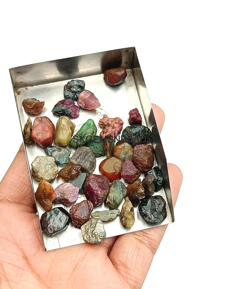 K&ouml;nnte beinhalten: Eine Sammlung roher, ungeschliffener Edelsteine in verschiedenen Farben, darunter Rot, Gr&uuml;n, Blau und Schwarz, die in einer kleinen Metallschale pr&auml;sentiert werden.