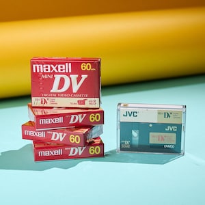 Op de afbeelding: Een stapel van vier rode Maxell Mini DV 60 digitale videocassettes met witte en gele tekst, naast een JVC Mini DV cassette in een doorzichtige behuizing. De achtergrond is geel en blauw.