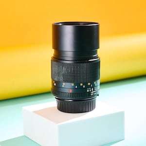 May include: A black camera lens with ribbed detailing and measurement markings. The lens is mounted on a white cube against a yellow and teal background. The lens has a black hood and a red dot.
