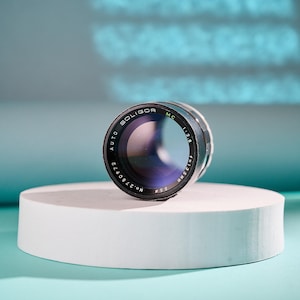 Puede incluir: Un objetivo de cámara Soligor Auto MC 1:2.8 f=135mm, negro con detalles plateados, se encuentra sobre una plataforma cilíndrica blanca. La lente tiene un elemento de vidrio teñido de azul sobre un fondo azul claro.