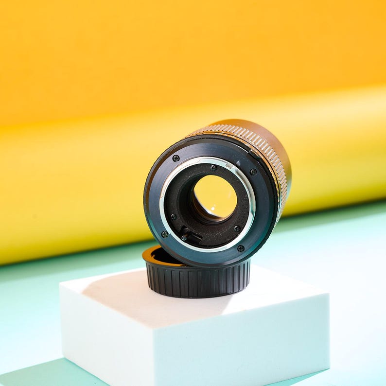 May include: A black and silver camera lens with a lens cap, set against a yellow and blue background. The lens is resting on a white cube. The lens has a circular opening and a ridged metal band.