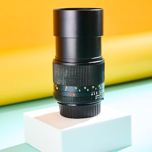 May include: A black camera lens with a ridged focus ring and measurement markings. The lens is cylindrical and sits on a white cube against a yellow and blue background. The lens has a black barrel and a series of numbers and markings.