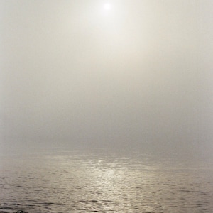 May include: A serene landscape photograph of a body of water under a hazy sky. The sun is a bright spot in the upper center. The water reflects the light, and the foreground shows a dark, textured shoreline.