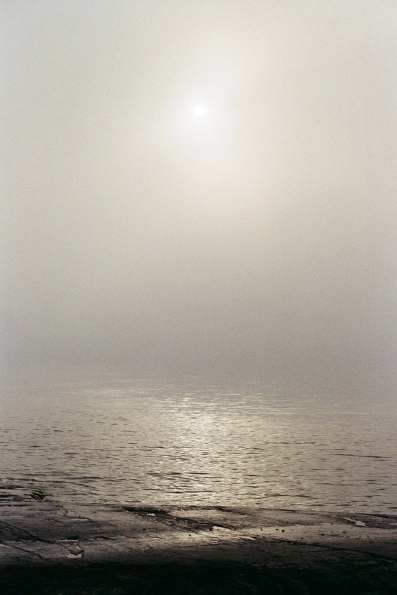 May include: A serene landscape photograph of a body of water under a hazy sky. The sun is a bright spot in the upper center. The water reflects the light, and the foreground shows a dark, textured shoreline.