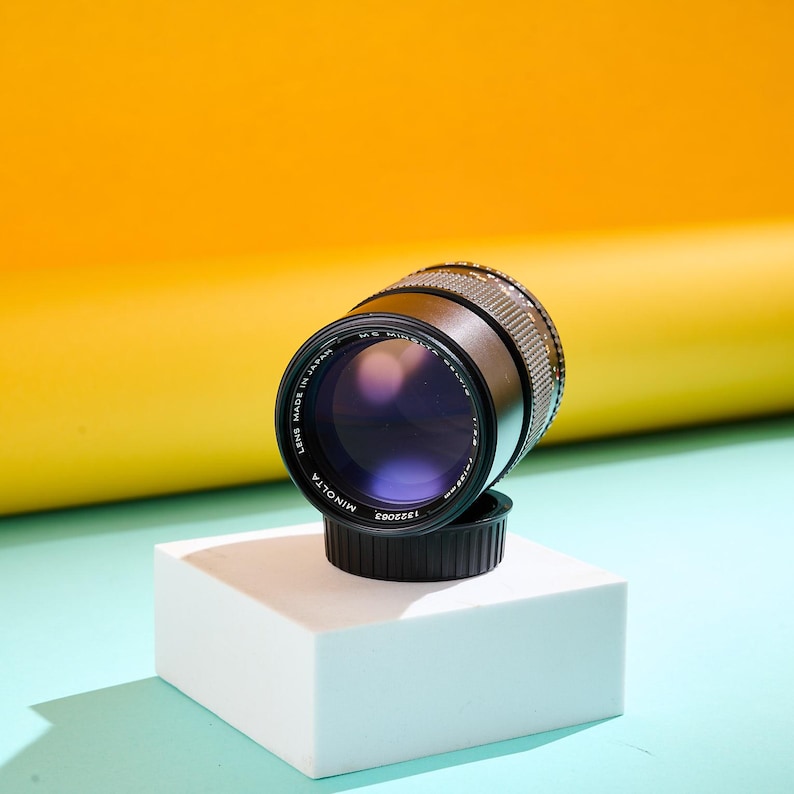 May include: A black Minolta camera lens with a silver textured grip, resting on a white cube. The lens has a purple hue and is set against a yellow and blue backdrop. The lens is labeled "Minolta" and "Made in Japan".