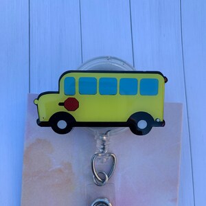 Può includere: Un porta badge a forma di scuolabus giallo con ruote nere, finestre blu e un segnale di stop rosso. Il porta badge ha un cordino retrattile in plastica trasparente e una clip in metallo. Lo sfondo è una carta a motivi rosa chiaro e bianco.