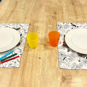 Peut inclure: Deux couverts avec des assiettes blanches, des verres transparents et des sets de table à colorier noir et blanc sur une table en bois. Les verres sont jaunes et orange. Il y a trois marqueurs de couleur sur le set de table de gauche.