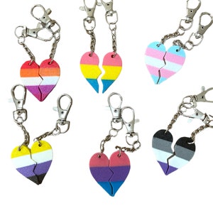 May include: Set of six split heart keychains featuring different pride flag colours. Each keychain has two halves of a heart, connected by a silver chain and a silver clip. The colours include: pink, orange, white, yellow, blue, green, light blue, dark blue, purple, black, white, and grey.