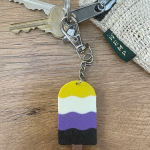 May include: A keychain with a yellow, white, purple, black, and brown popsicle charm. The charm is shaped like a popsicle and has the colors of the non-binary pride flag. The keychain is attached to a silver key ring and a silver chain.