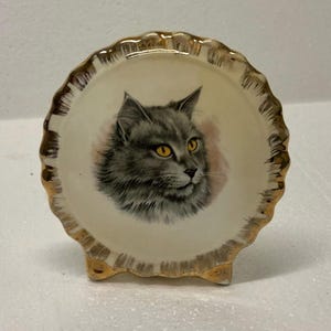 May include: A small, decorative ceramic dish with a scalloped edge and three small feet. The dish features a detailed illustration of a gray cat with yellow eyes. The rim is trimmed with gold paint, adding a touch of elegance.