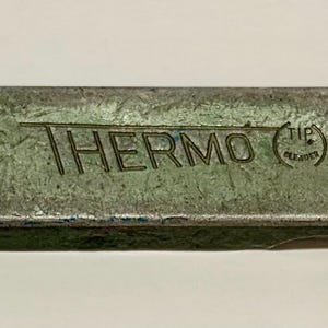 May include: A rectangular, vintage metal object with the word "THERMO" in dark lettering. The object has a distressed, green-gray finish and a small circular detail on one end. The text "TIP CLEANER" is also visible.