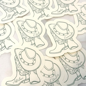 May include: A collection of stickers featuring a cowboy hat and boots design. Each sticker has a name written on it, such as "Allie", "Steven", "Chad", "Leonard", "Adeline", "Charles", "Jason", and "Sophia". The stickers are outlined in green.