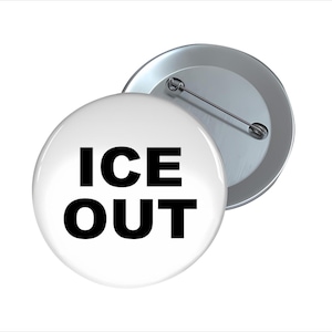 May include: A white button with the words "ICE OUT" in bold black letters. The button has a shiny, reflective surface and a metal pin backing. Another button is partially visible.