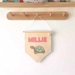 May include: A beige felt pennant with the name "Millie" embroidered in pink letters and a green turtle illustration. The pennant is hanging on a wooden shelf with hooks, above two children's books and a grey elephant soft toy.