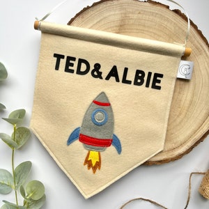 May include: A beige felt banner with the names "TED & ALBIE" in black lettering. A grey felt rocket with red and blue accents is centred on the banner. The banner is hanging from a wooden dowel.