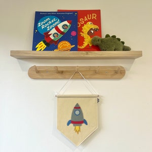 May include: A beige felt banner with a grey rocket ship with red and blue accents. The banner is hanging from a wooden shelf with two books and a green stuffed dinosaur on top.