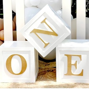 May include: Three white blocks with gold letters spelling out "ONE". The blocks are stacked on top of each other.