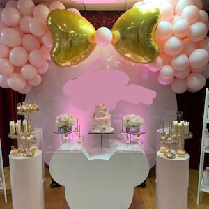 May include: A white Mickey Mouse-shaped table with a clear glass top. The table is decorated with pink and gold balloons, a pink and white cake, and floral arrangements. There are also two white pedestals with gold accents.