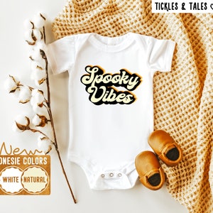 Op de afbeelding: Witte baby romper met de tekst "Spooky Vibes" in een retro lettertype met gele en zwarte accenten. De romper ligt op een witte achtergrond met een bruin gebreid deken en katoenen bollen.