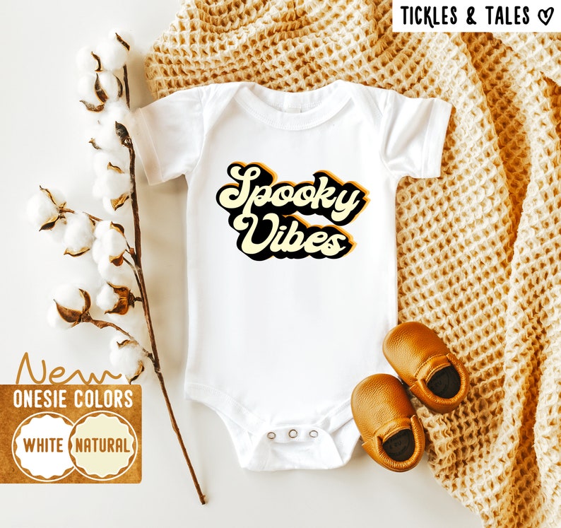 Op de afbeelding: Witte baby romper met de tekst "Spooky Vibes" in een retro lettertype met gele en zwarte accenten. De romper ligt op een witte achtergrond met een bruin gebreid deken en katoenen bollen.