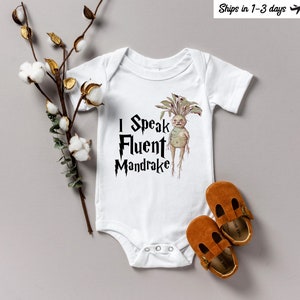 Könnte beinhalten: Weißer Baby-Body mit dem Text "I Speak Fluent Mandrake" und einer Mandrake-Illustration. Zwei braune Babyschuhe aus Leder liegen neben dem Body. Ein Zweig mit Baumwollstielen befindet sich auf der linken Seite des Bildes.