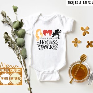 Pode incluir: Macacão branco com um gráfico de três bruxas com o texto "It's all a bunch of Hocus Pocus".