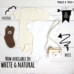 Op de afbeelding: Twee baby rompers, een witte en een naturel, met een houten speelgoedcamera en een paar bruine sokken met een berenkop. De tekst "Now Available in White & Natural" is onder de rompers gedrukt. De tekst "See Sizes" is gedrukt op een vlag met een pijl die naar rechts wijst.