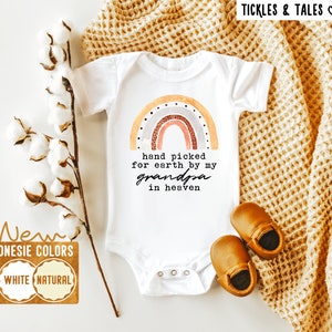 Peut inclure: Combinaison blanche avec un motif arc-en-ciel et le texte "hand picked for earth by my grandpa in heaven". La combinaison est sur un fond blanc avec une couverture marron et des fleurs de coton.