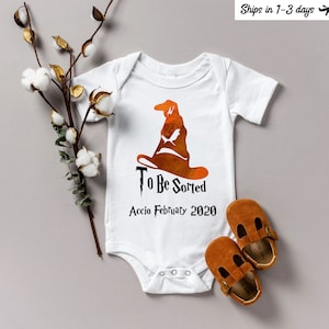 Può includere: Body da bambino bianco con una grafica del Cappello Parlante e la scritta "To Be Sorted Accio February 2020". Due scarpine da bambino in pelle marrone sono accanto al body. Un rametto di cotone ed eucalipto è sul lato sinistro dell'immagine.