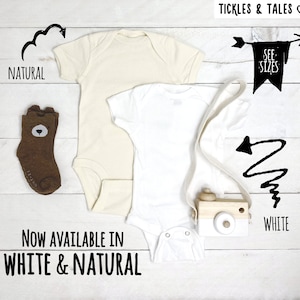 May include: Two baby onesies, one white and one natural, with a wooden toy camera and a pair of brown socks with a bear face. The onesies are available in white and natural. "See Sizes" is written on a banner.