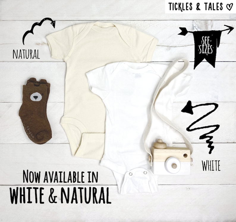 May include: Two baby onesies, one white and one natural, with a wooden toy camera and a pair of brown socks with a bear face. The onesies are available in white and natural. "See Sizes" is written on a banner.