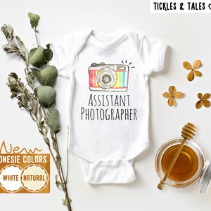 Może przedstawiać: Biały śpioszek dla niemowlęcia z kolorową kreskówkową kamerą i tekstem "Asystent fotografa".