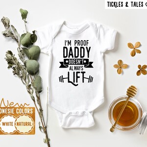May include: White baby onesie with black text that reads "I'm Proof Daddy Doesn't Always Lift". The onesie has a white background and black text. The text is in a bold, sans-serif font. The onesie is on a white background.
