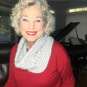 May include: A woman with curly blonde hair is wearing a red sweater and a white fuzzy scarf. She is sitting in a chair in front of a black grand piano.