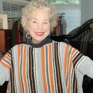 May include: A handmade crochet poncho with vertical stripes in orange, black, white, and gray. The poncho has short sleeves and is worn over a black turtleneck. The woman is smiling and wearing hoop earrings.