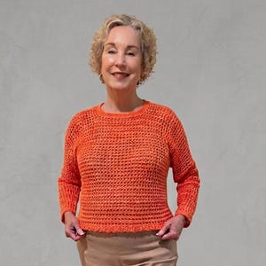 Puede incluir: Un suéter naranja de crochet con cuello redondo y mangas largas. El suéter tiene un diseño texturizado y de tejido abierto. La persona lleva pantalones color beige. El fondo es de color gris claro liso.