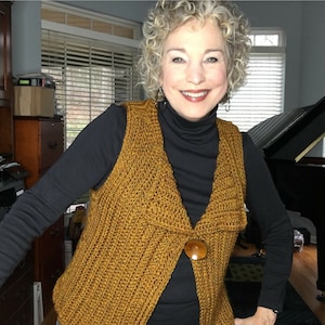 May include: A woman wearing a brown crocheted vest with a large brown button. She is wearing a black turtleneck shirt and has curly gray hair.