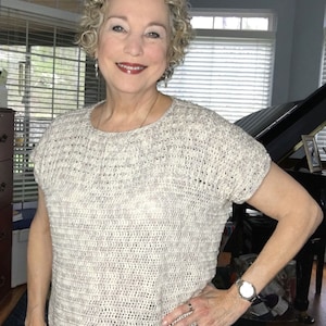 May include: A woman wearing a white crocheted top with a loose fit and short sleeves. The top has a textured pattern and is made of a soft, lightweight yarn.
