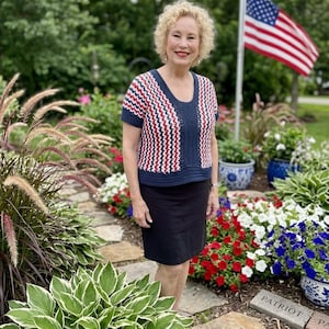 May include: A crochet top with a red, white, and blue chevron pattern. The top has short sleeves and a scoop neck. The woman is wearing a black skirt and standing in a garden with an American flag in the background.
