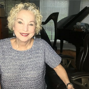 Pode incluir: Uma mulher usando um top de crochê azul e cinza de manga curta. Ela está sentada em uma cadeira de escritório preta, com um piano de cauda e uma harpa visíveis ao fundo. A mulher tem cabelo loiro encaracolado e está sorrindo.