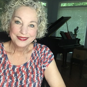 May include: A woman with curly grey hair is wearing a red, white and blue patterned shirt. She is sitting in a chair in front of a black grand piano.