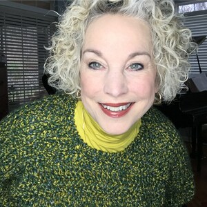 May include: A woman with curly, gray hair is wearing a green and yellow sweater. She is smiling and looking directly at the camera.