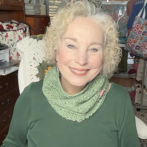 May include: A woman wearing a green crocheted cowl neck warmer. The cowl is a light green color and has a textured stitch pattern. The woman is also wearing a green long-sleeved shirt.