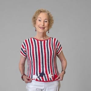 May include: A short-sleeved crochet top featuring vertical stripes in red, white, and navy blue. The hem is finished with a decorative scalloped edge, small white floral accents, and a navy blue star-shaped detail centred at the bottom.